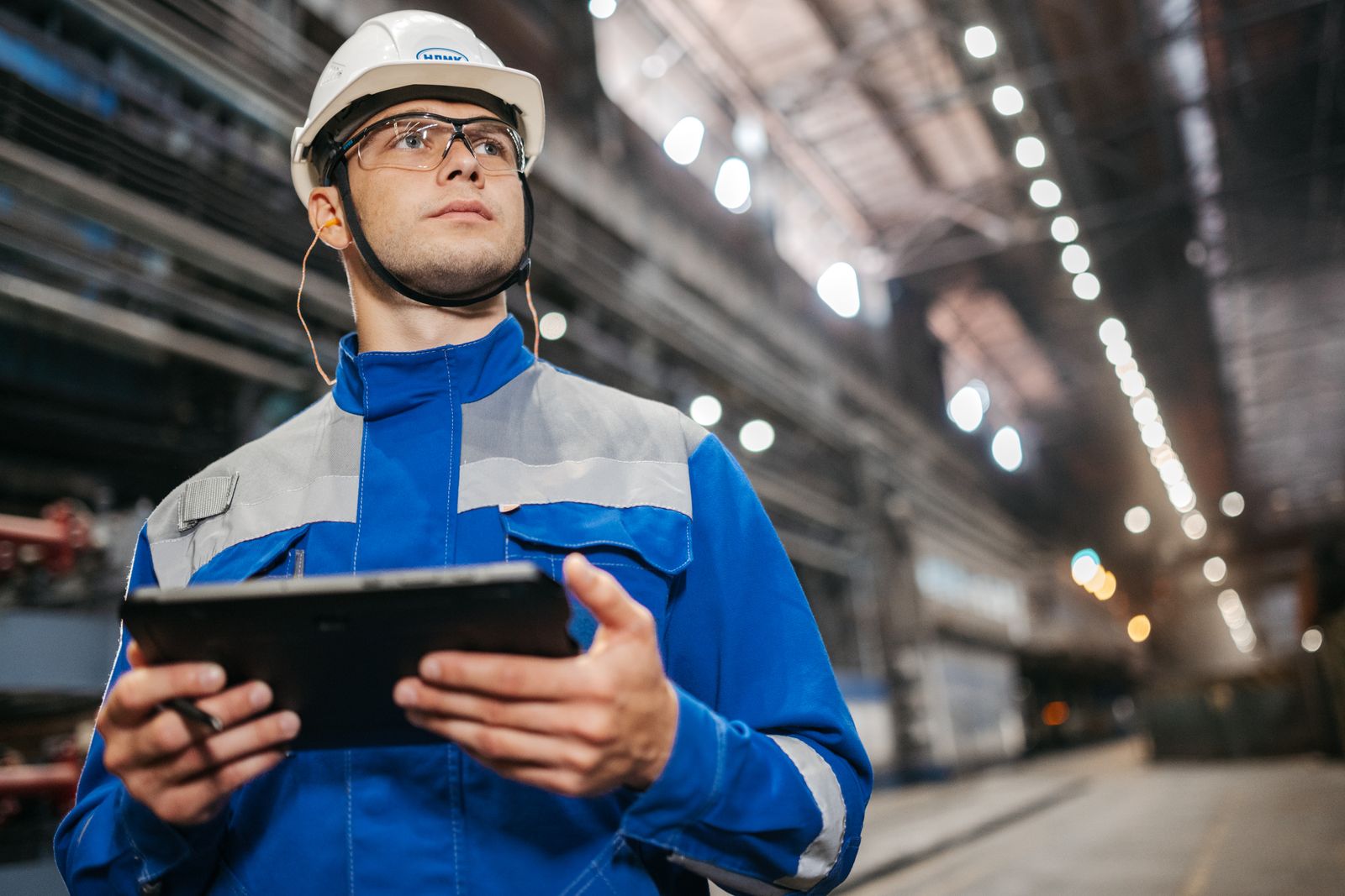 Two industrial workers at a plant reviewing a digital permit on a tablet before starting hot work, hi-vis and hard hats, an isolation point visible behind them.